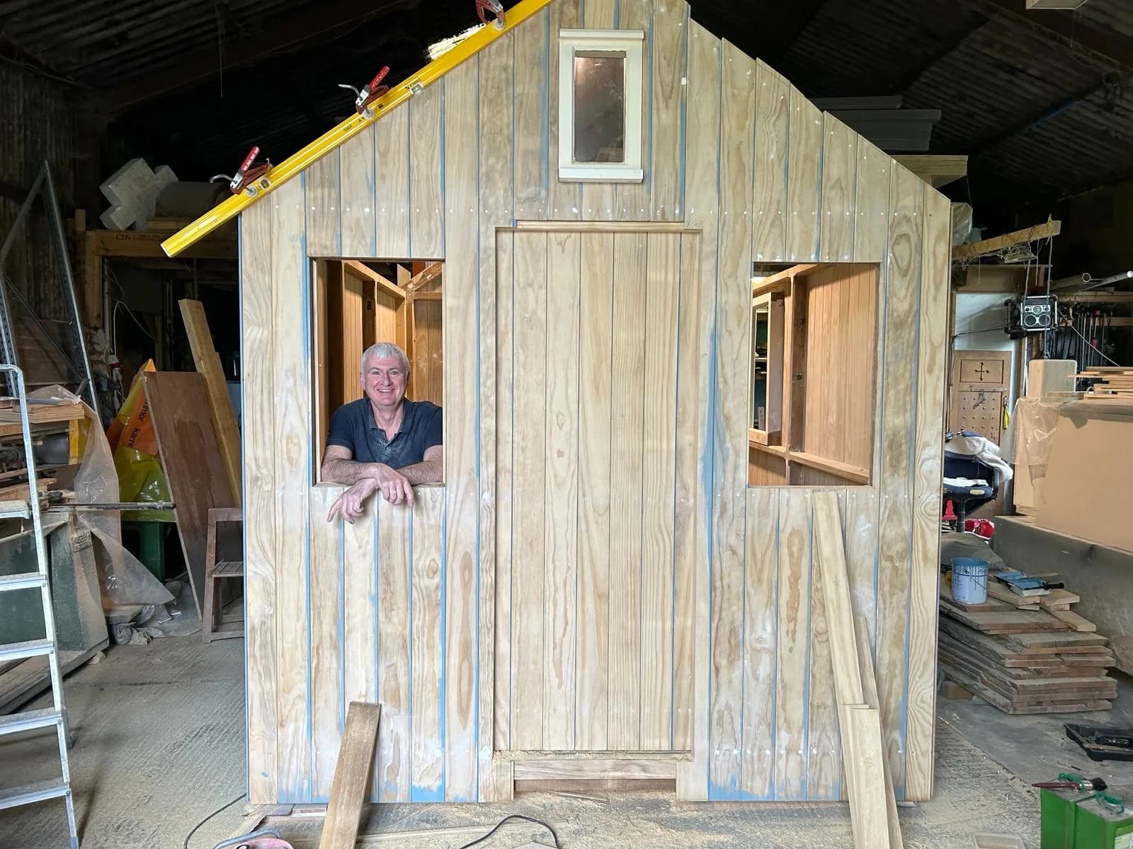 Neil smiling through the window opening of a timber-clad bespoke shed being built in the workshop